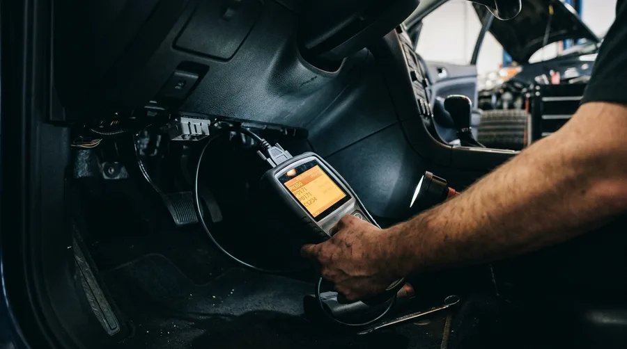 a scan tool plugged into an OBD port under a dashboard, screen glowing with diagnostic codes, low ambient light in a shop bay